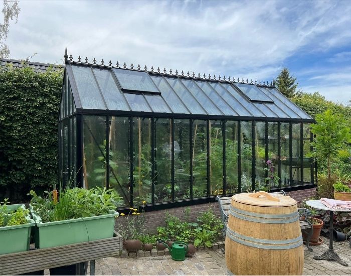 Impressie van een grote tuinkas in een royale tuin. De tuinkas heeft veiligheidsglas, een sterk aluminium frame in de kleur zwart en klassieke Victoriaanse nok.